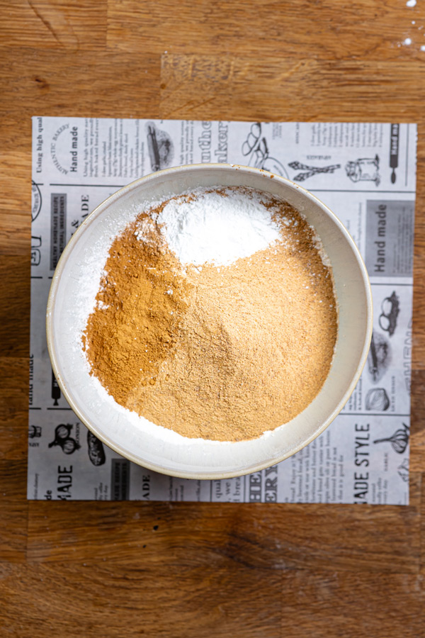 trockene zutaten fuer glutenfreier apfelkuchen von oben fotografiert in einer schuessel auf einem holztisch mit weissem backpapier