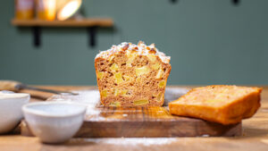 angeschnittener apfelkuchen mit dinkelmehl aus der kastenform frontal auf einem holzbrett mit dekoration