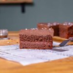 ein stueck vegane schokotorte von der seite mit sichtbaren schichten und uebriger torte im hintergrund
