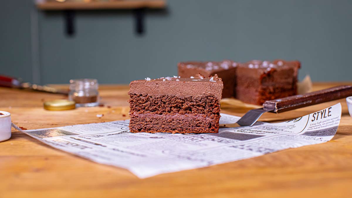 ein stueck vegane schokotorte von der seite mit sichtbaren schichten und uebriger torte im hintergrund