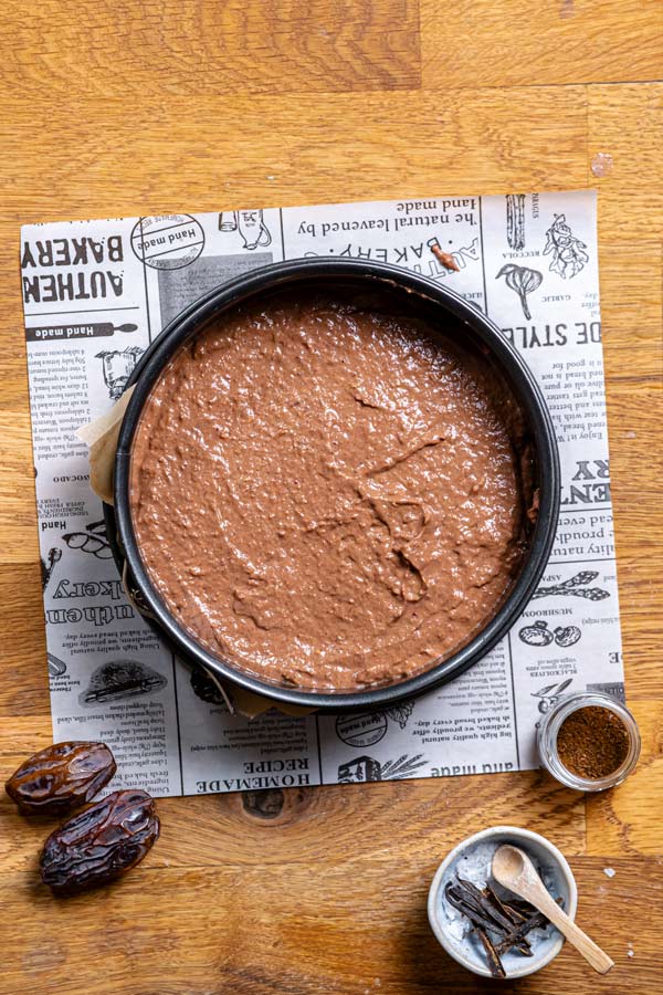 teig fuer vegane schokotorte in einer runden schwarzen springform auf einem holztisch mit weissem backpapier