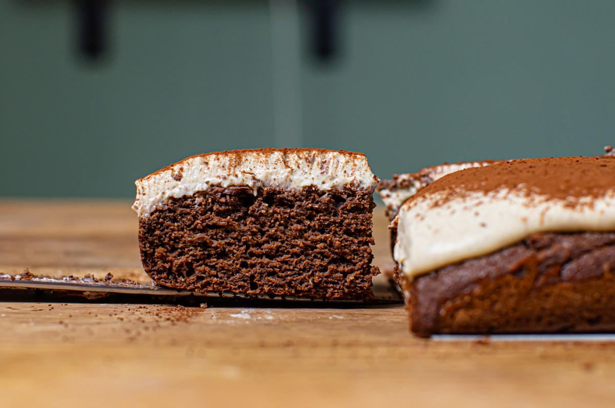 herausgeschnittenes stueck schoko protein kuchen mit mascarpone frontal auf einem holztisch mit uebrigem kuchen am rand