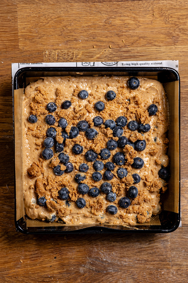 teig und streusel sowie heidelbeeren in einer schwarzen quadratischen backform von oben auf einem holztisch