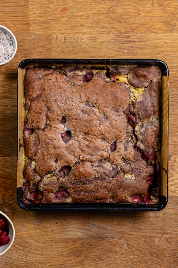 gebackener erdbeer schokokuchen mit cheesecake fuellung in einer quadratischen schwarzen backform von oben auf einem holztisch