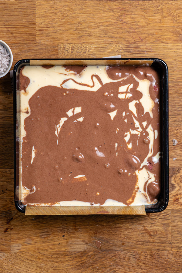 alle schichten fuer erdbeer schokokuchen mit cheesecake fuellung in einer schwarzen quadratischen backform von oben auf einem holztisch