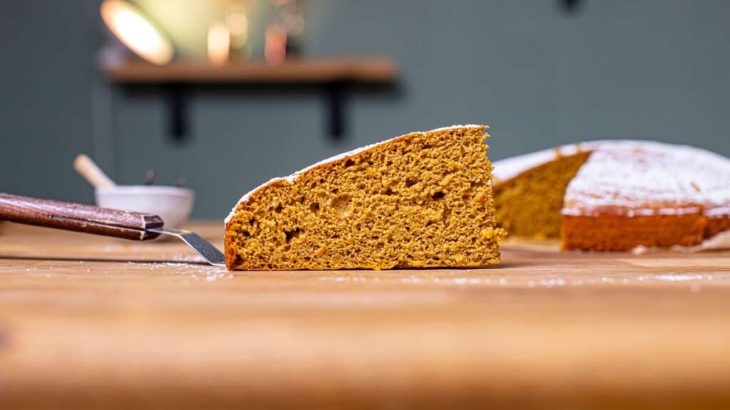 ein stueck moehrenkuchen von der seite auf einem holztisch mit dem restlichen kuchen im hintergrund