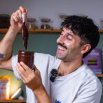 mann haelt ein glas mit selbstgemachter dattel haselnuss creme und laesst die schokoladige masse vom loeffel zurueck ins glas fliessen