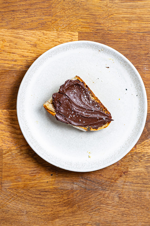 eine halbe mit dattel haselnuss creme bestrichene brotscheibe auf einem teller auf einem holztisch von oben
