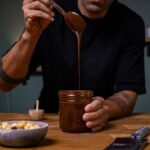 maennliche person haelt nutella alternative im glas auf holztisch von vorne