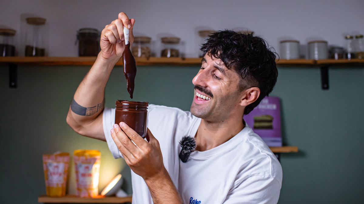 mann haelt ein glas mit selbstgemachter dattel haselnuss creme und laesst die schokoladige masse vom loeffel zurueck ins glas fliessen