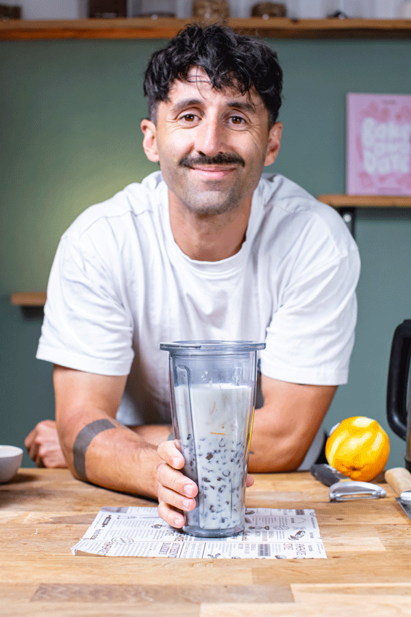 braunhaariger mann mit schnurbart lehnt auf einem holztisch und haelt einen mit milch gefuellten mixbehaelter in der rechten hand