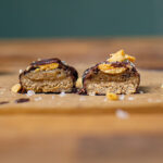 selbstgemachtes gesundes snickers halbiert mit beiden haelften nebeneinander auf einem holzbrett von vorne