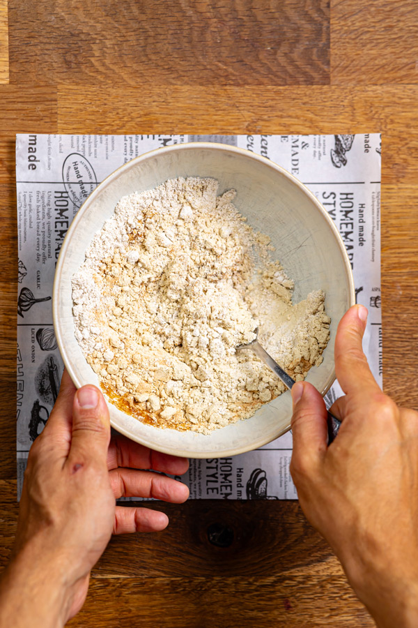 trockene zutaten fuer gesundes snickers in einer schuessel von oben auf weissem backpapier auf einem holztisch mit zwei haenden