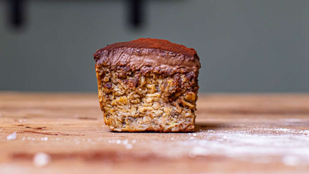 ein stueck baked oats mit banane mit frosting von vorne auf einer holzplatte