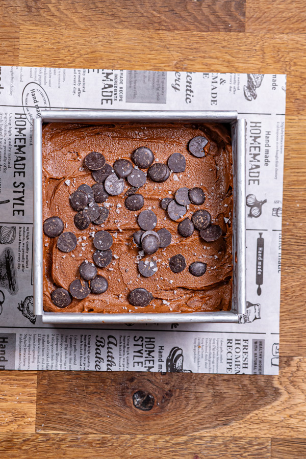 brownie ohne mehl masse in einer quadratischen backform mit schokodrops garniert von oben auf einem holztisch