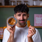 mann haelt schmunzelnd mixbehaelter mit lotus creme alternative und lotus keks in die kamera