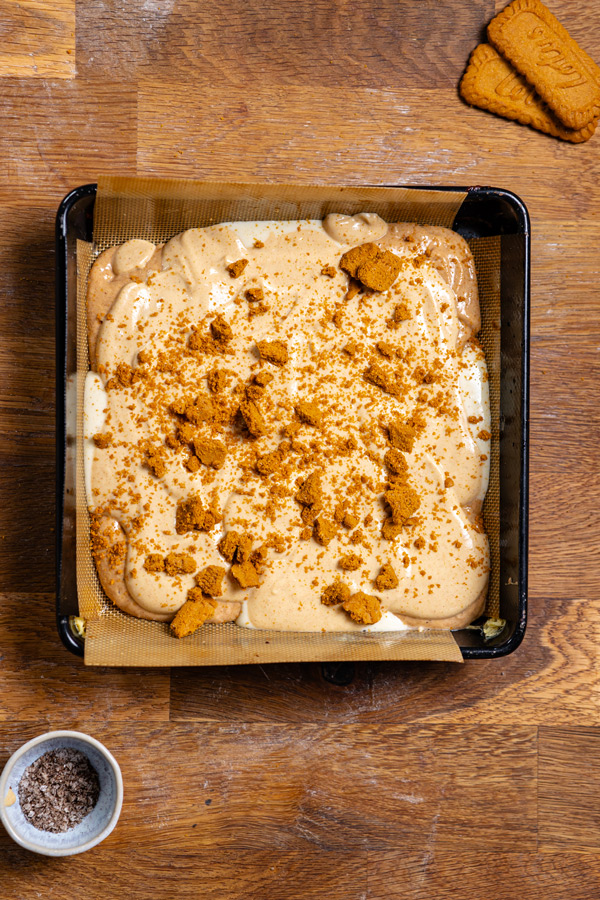 pudding und lotus keks schicht fuer biscoff kuchen in einer schwarzen quadratischen backform von oben auf einem holztisch